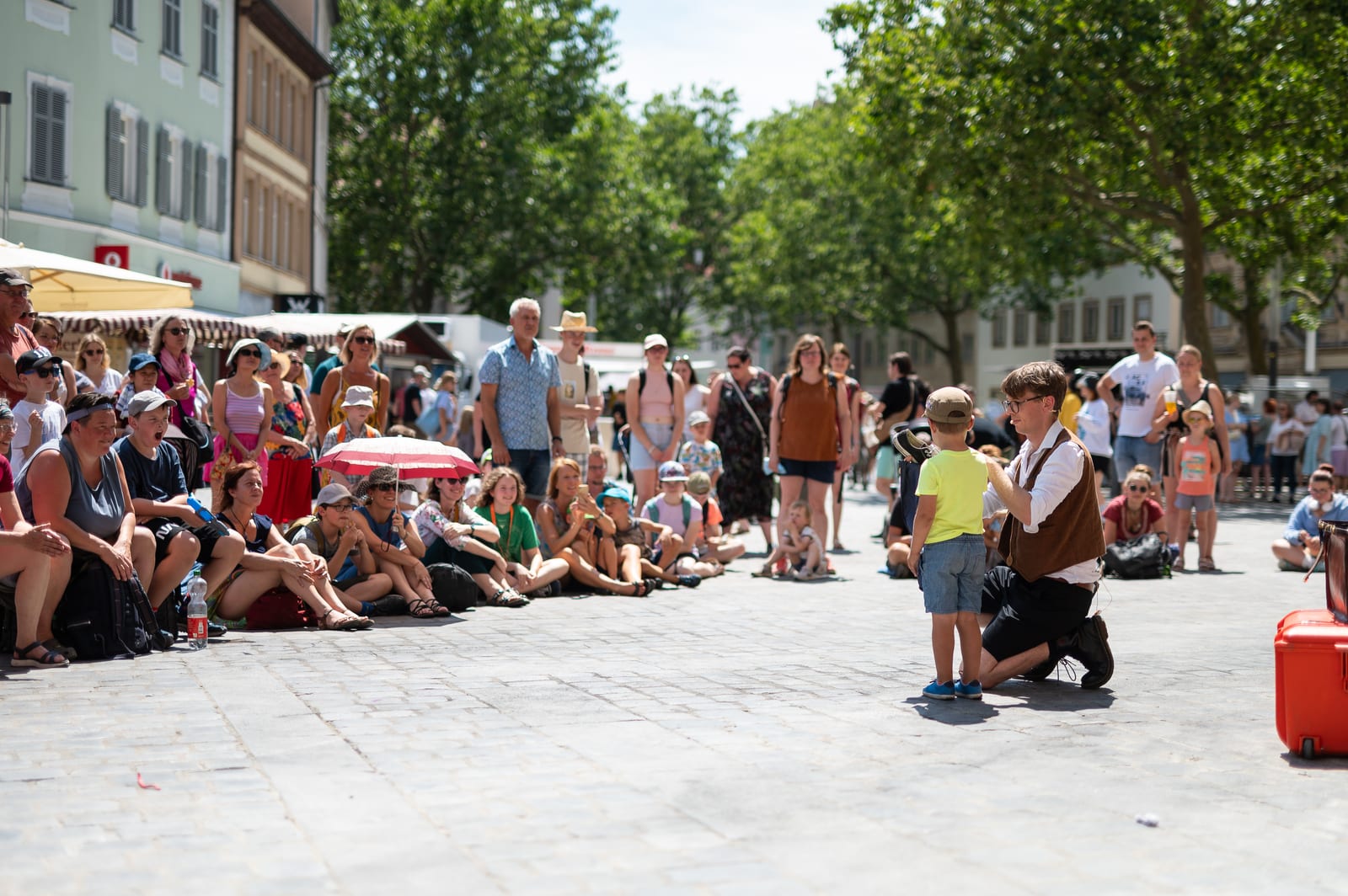Audience seated in a semicircle on the ground watching Sam King perform with a young volunteer at the Bamberg Magic Festival