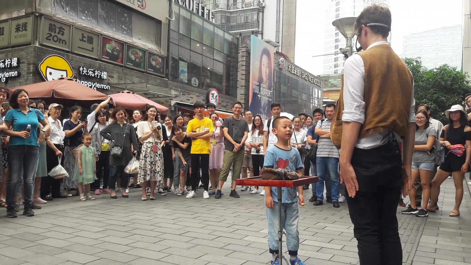 A standing crowd watching Sam King perform magic with a young volunteer at a table on a street in Shanghai
